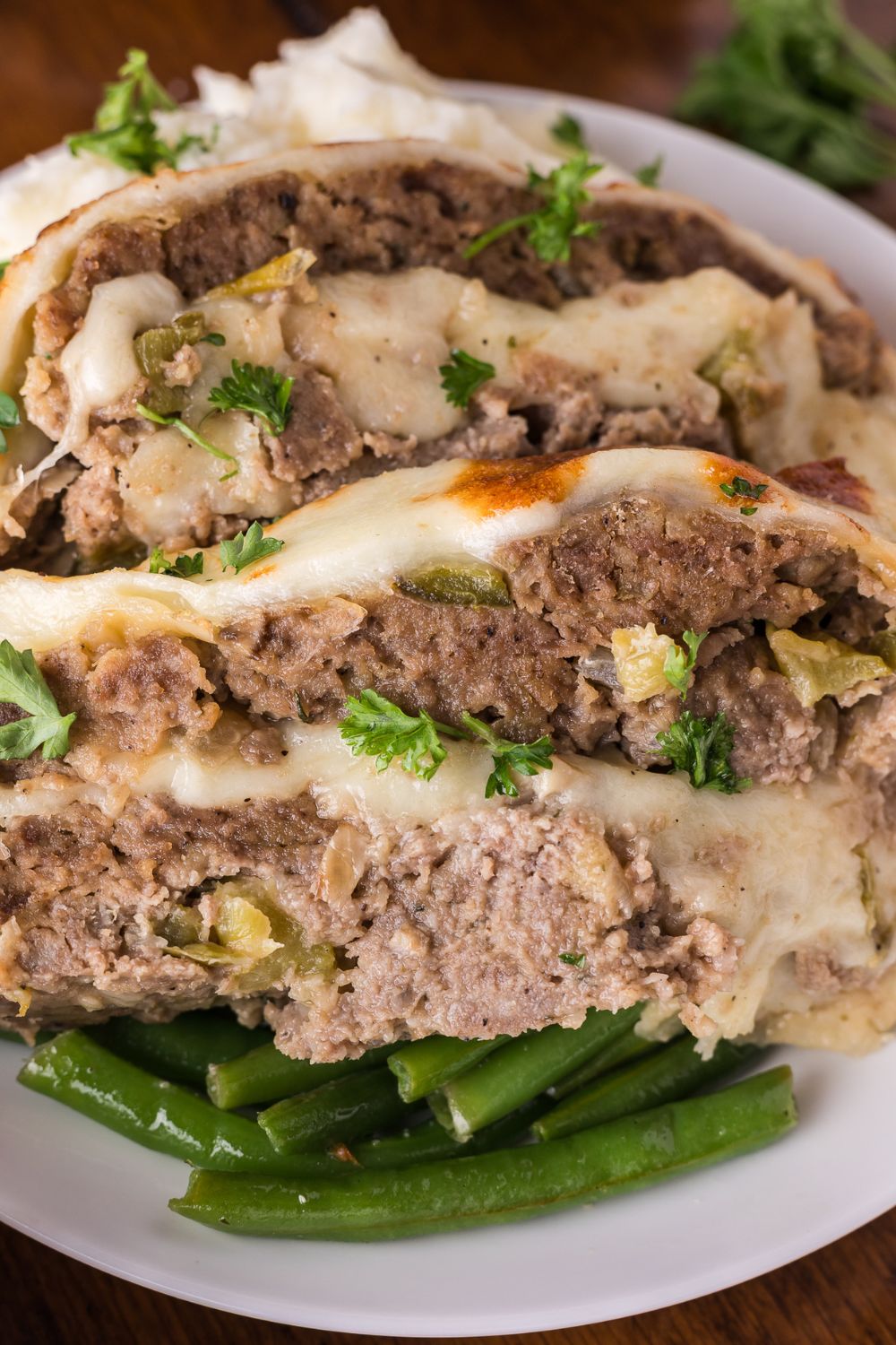 white plate sitting on table with a bed of green beans, and on top thick slices of Stuffed Philly Cheesesteak Meatloaf topped with fresh parsley