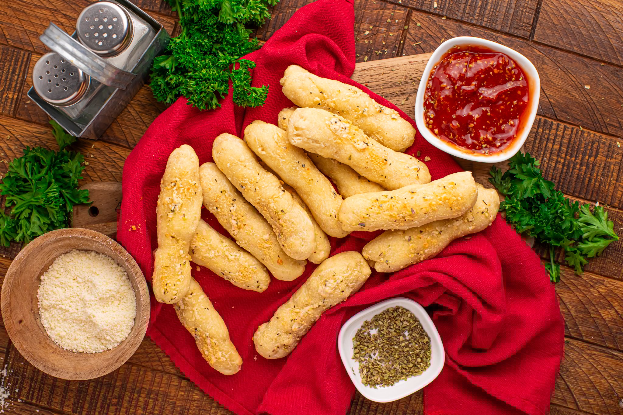 overhead look of breadsticks with small bowls of sauce, parmesan cheese & oregano