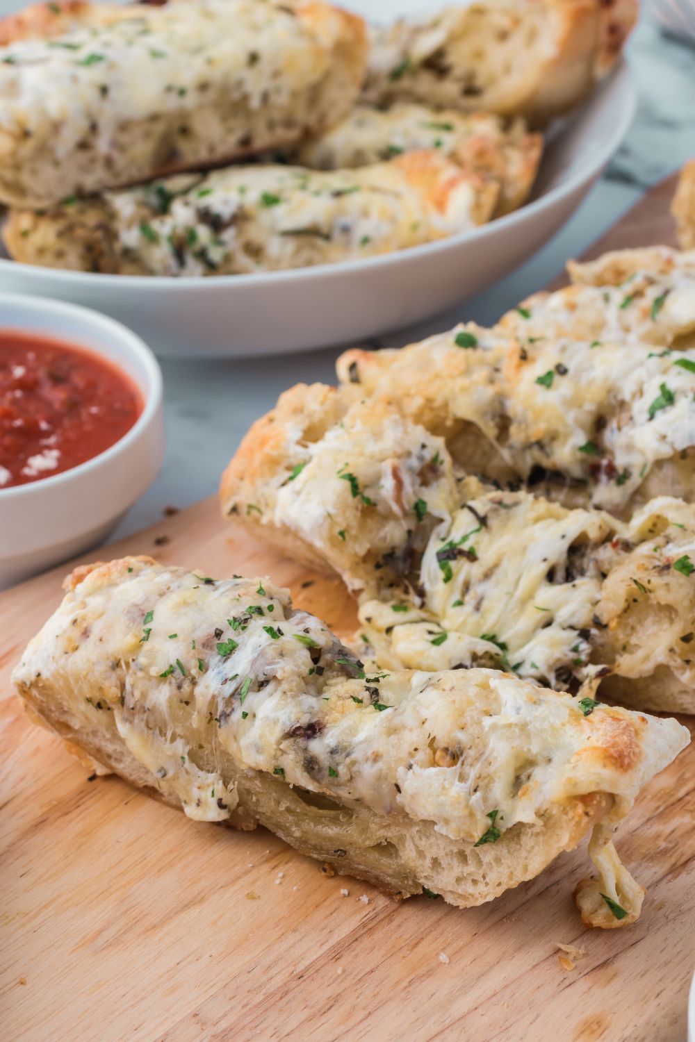 Cheesy Garlic Bread sliced and placed on table with marinara in bowl and more breadsticks in a bowl behind it on table 
