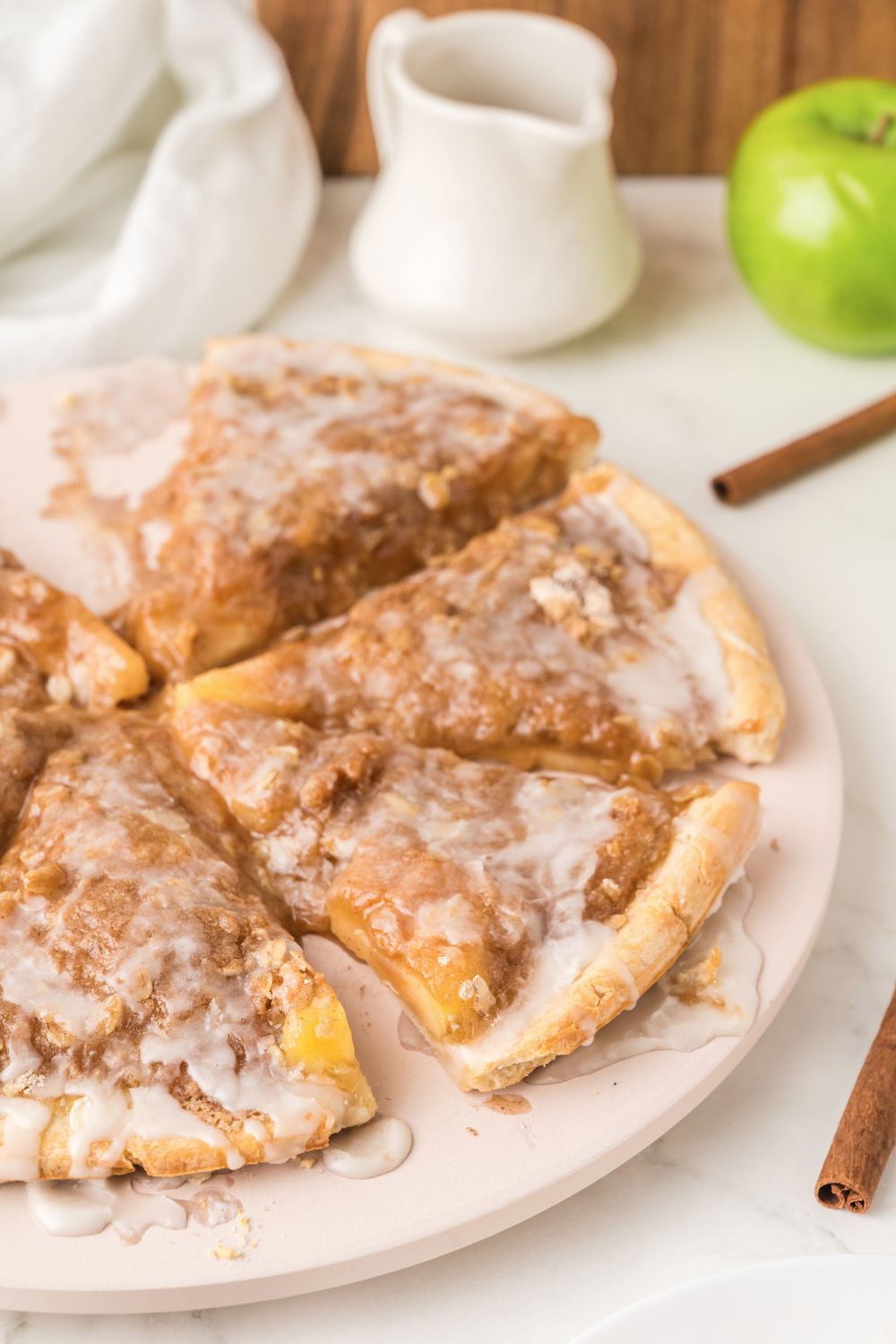 side view of Apple Pie Pizza on table with some cinnamon sticks to side and a green apple to side 