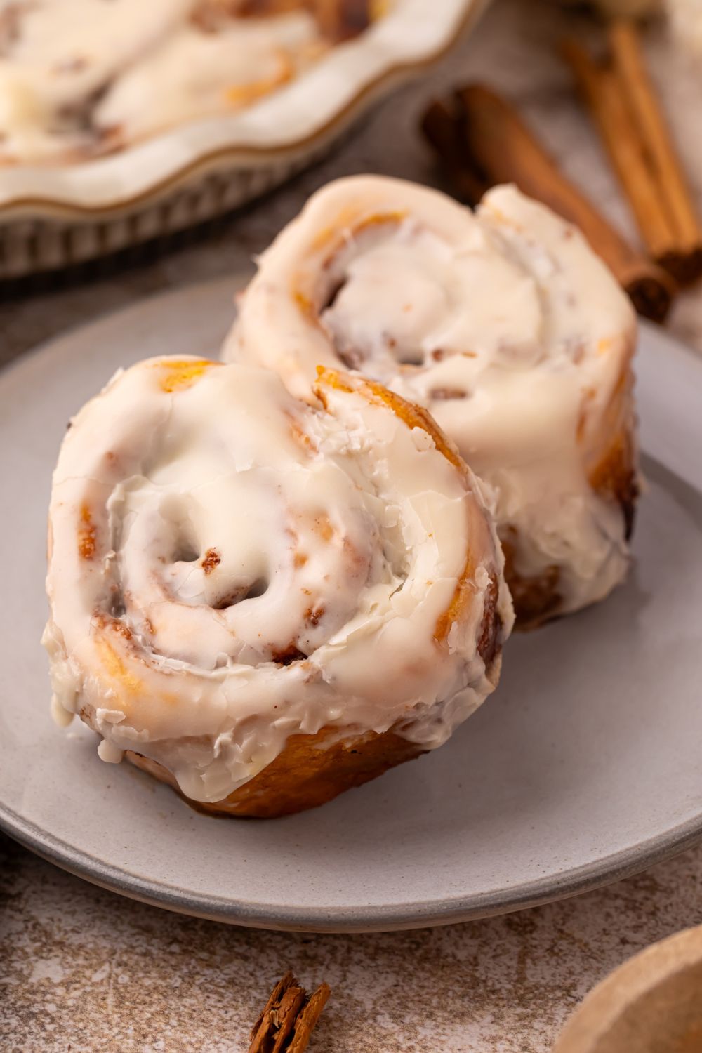 2 Pumpkin Cinnamon Rolls on a grey plate on table with a pan with more rolls behind it and a couple of cinnamon sticks on table 