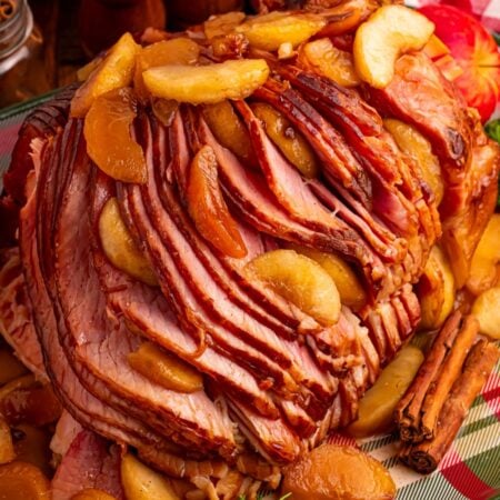 Slow Cooker Apple Glazed Ham on a plaid platter sitting on table with a checkered red towel behind ham, salt and pepper shakers
