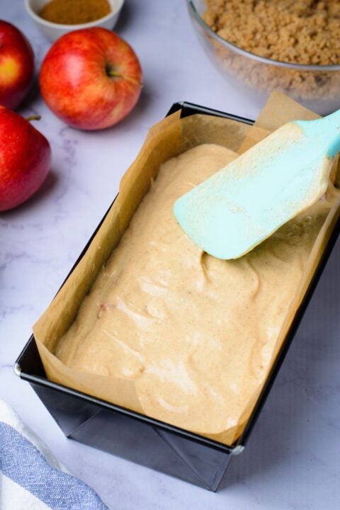 raw batter in a loaf pan being spread around with a spatula, and two apples to side of pan 