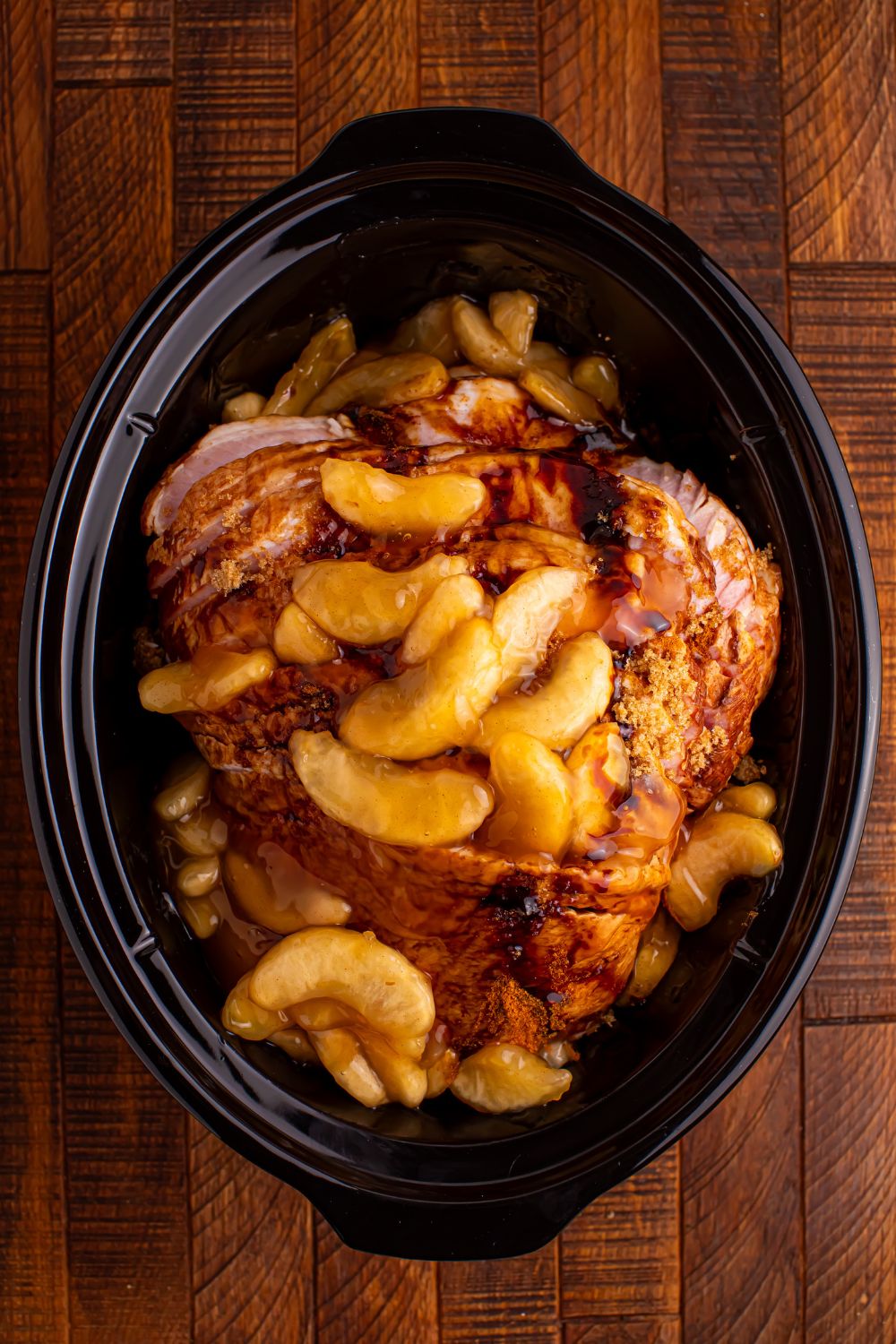 wooden table with a black crockpot in center with a sliced ham topped with spiced apples 