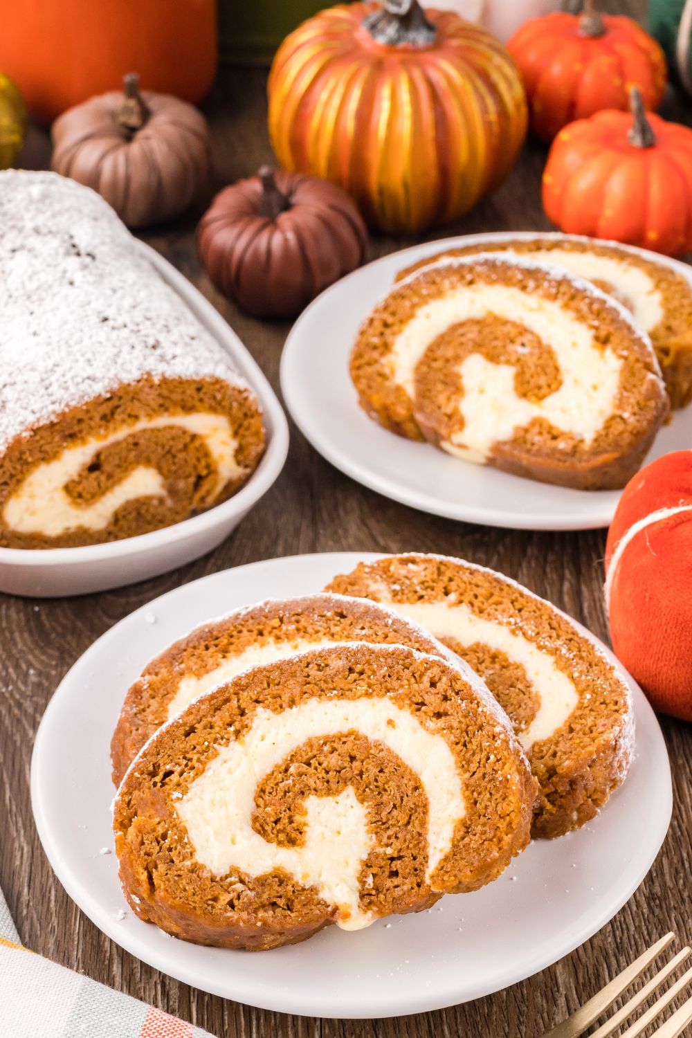 Pumpkin Cake Roll placed on plates, with a small platter to side with more of the pumpkin roll. Mini pumpkins scattered on table for a Thanksgiving fall look 