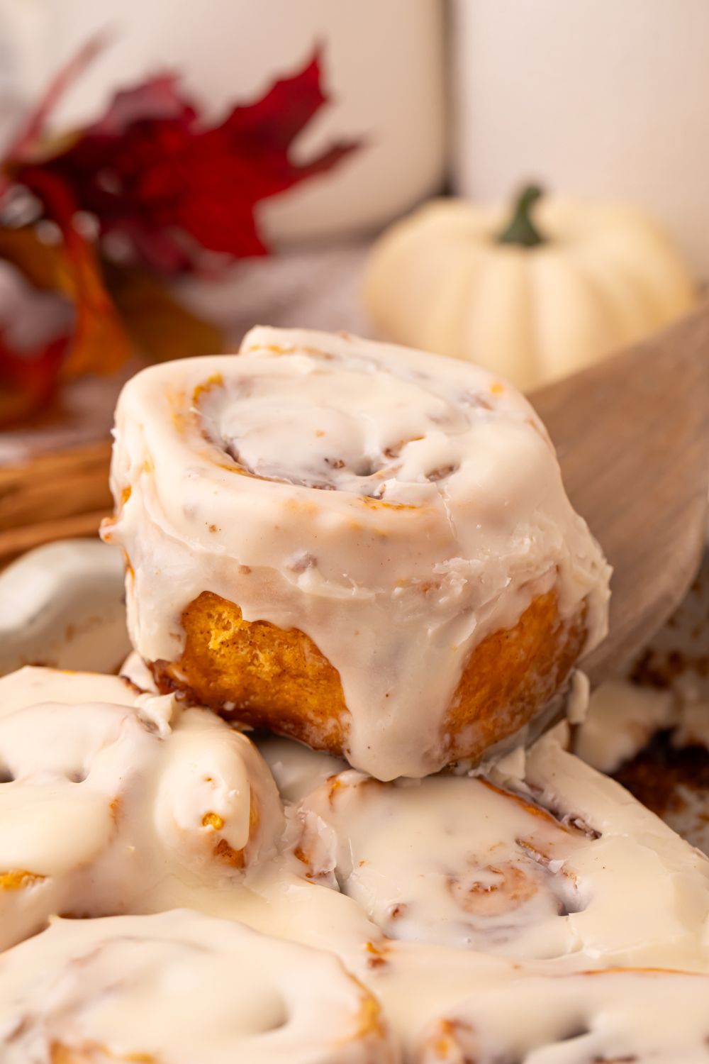 Pumpkin Cinnamon Roll being lifted out of container with a wooden spoon. The back of the image is decorated with fall items like leaves, pumpkins, etc