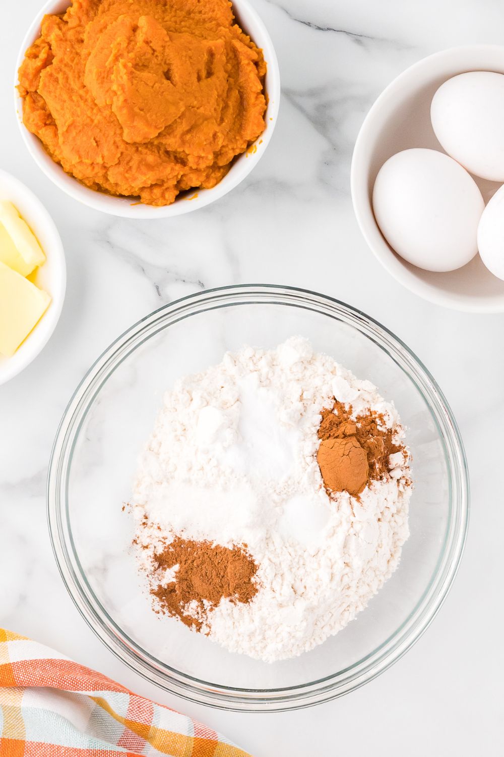 flour, spices and more in a glass bowl with small bowls around it with eggs, pumpkin and sliced butter for making Pumpkin Cake Roll