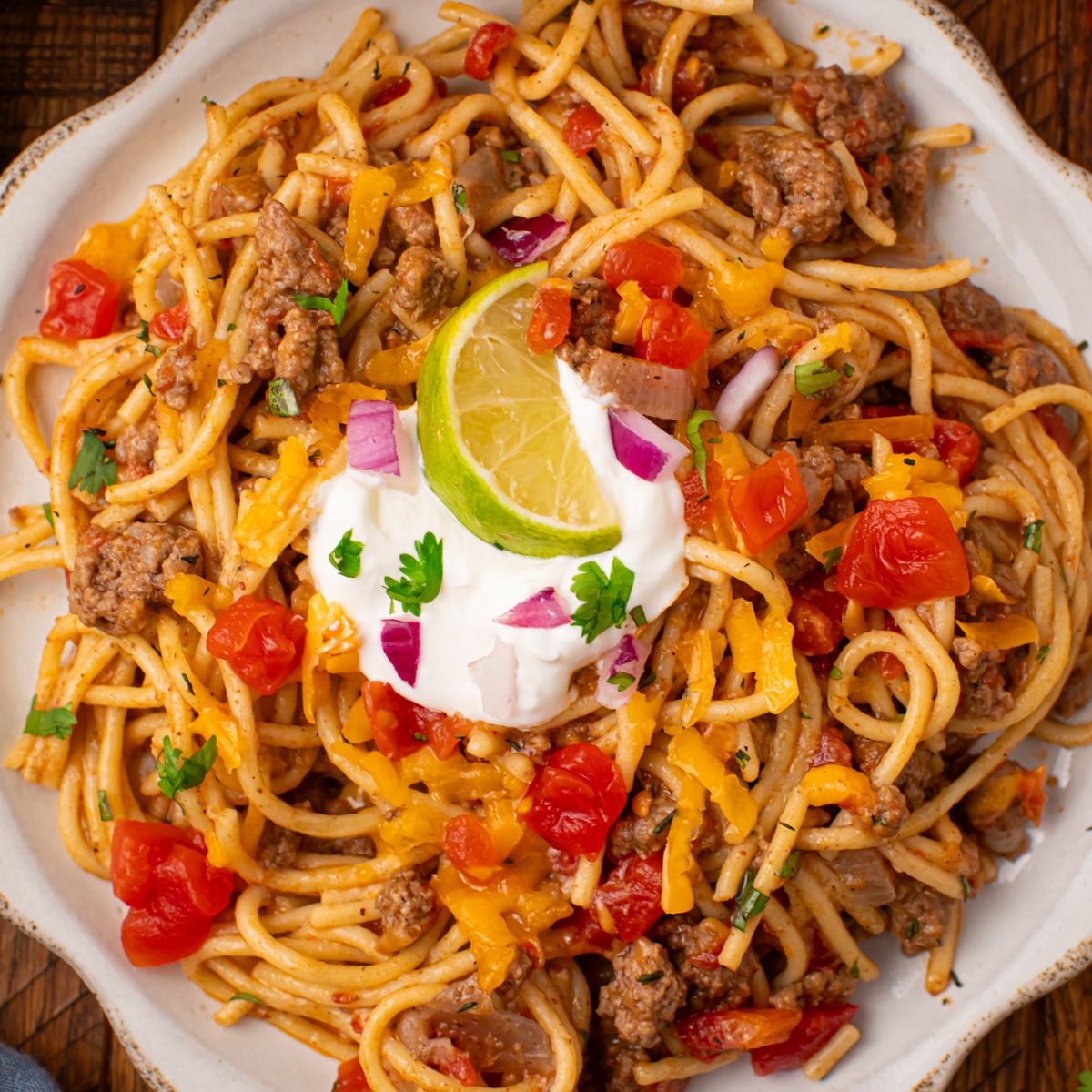 white plate on wooden table that is piled full of Cheesy Taco Spaghetti with sour cream and lime wedge on top of mixture