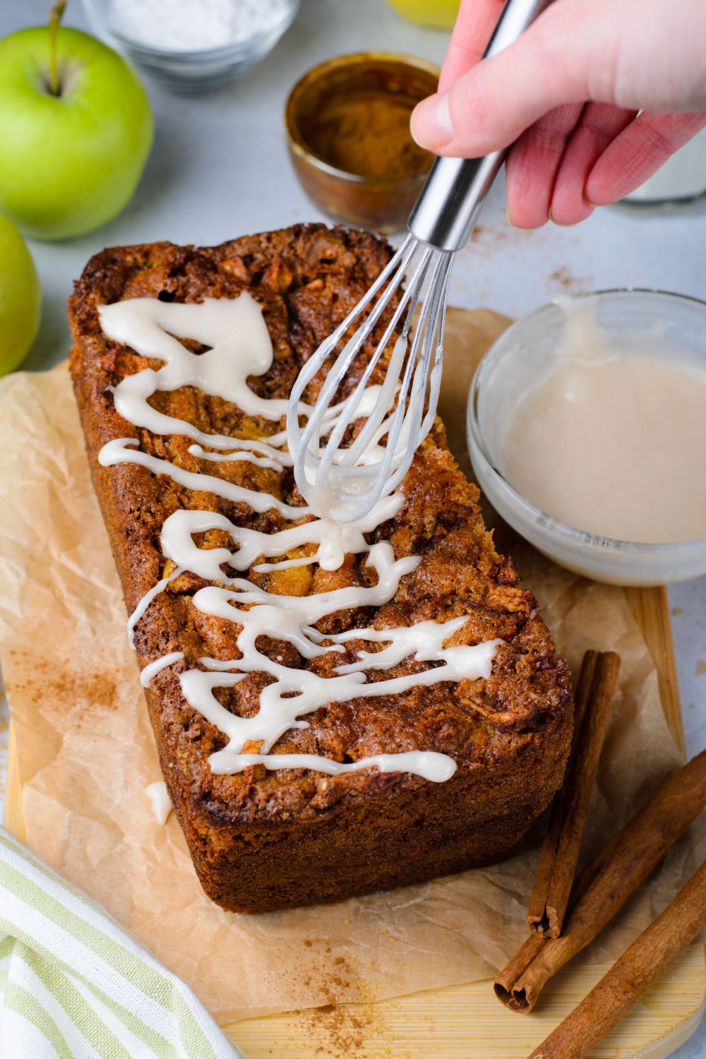 amish apple bread on table with a whisk drizzling glaze over the top with a small bowl of glaze to side 