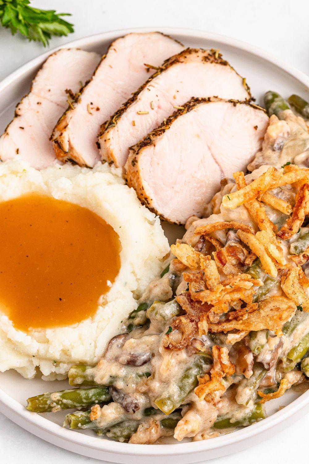 a white plate on a table with slices of turkey, scoop of mashed potatoes and gravy and some green bean casserole on it 