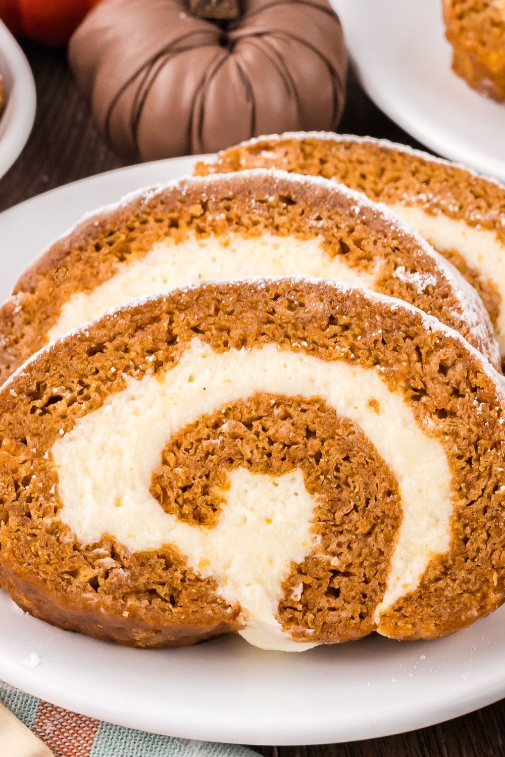 a white plate on table with three thick slices of Pumpkin Cake Roll on it, and mini pumpkins to side of plate 