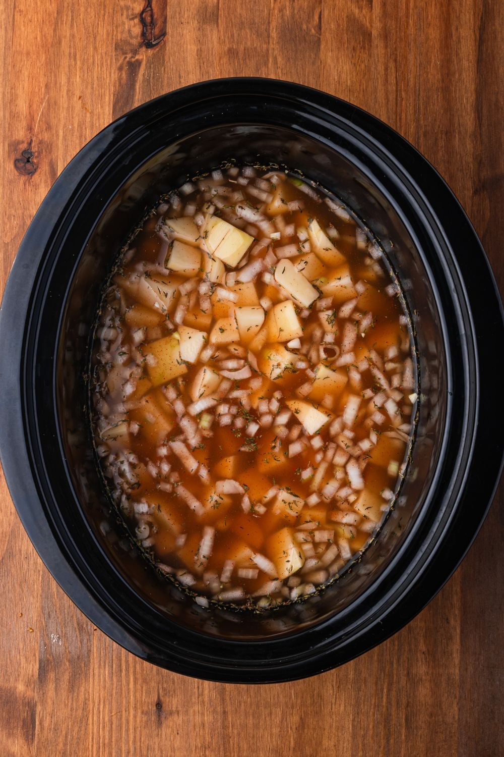 black crockpot sitting on a wooden  table with diced potatoes, onions, beef broth in more in the slow cooker