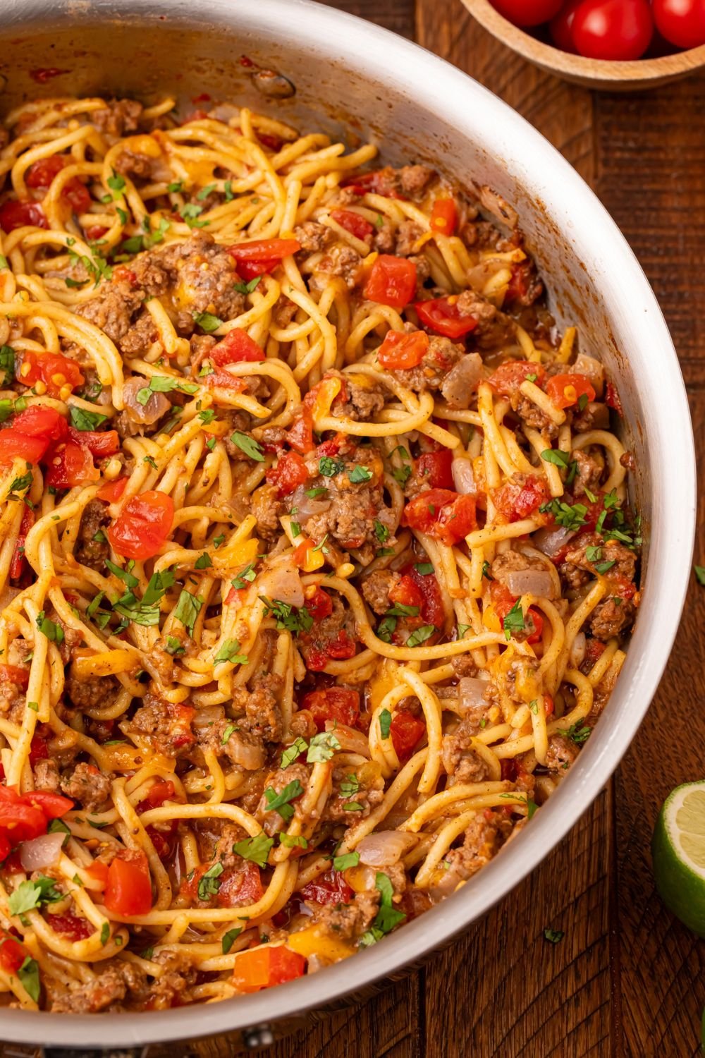 wooden table with a metal skillet full of Cheesy Taco Spaghetti in pan and a small bowl of tomatoes to side