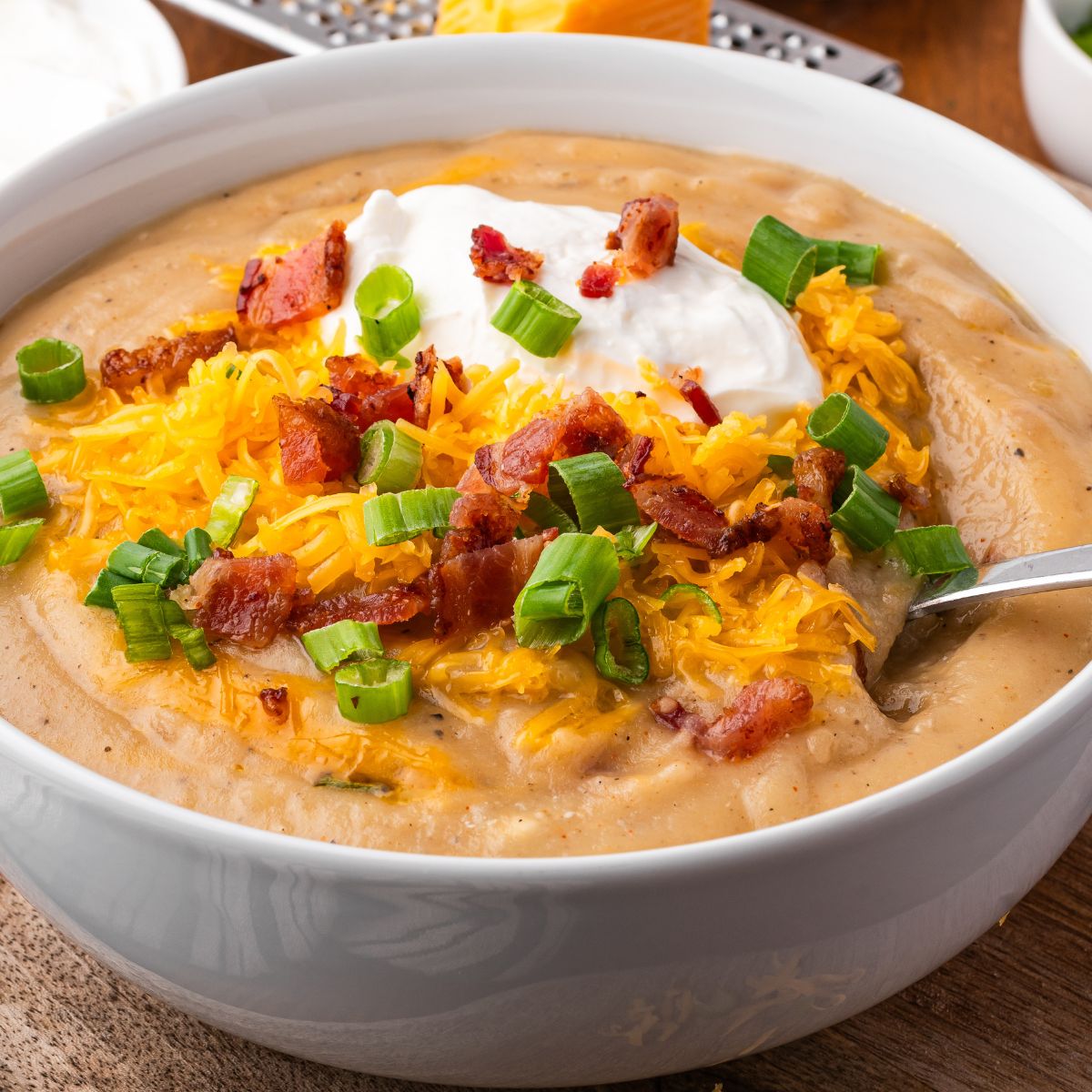 wooden table with a white bowl full of Crockpot Loaded Baked Potato Soup topped with shredded cheese, bacon crumbles, green onions and sour cream