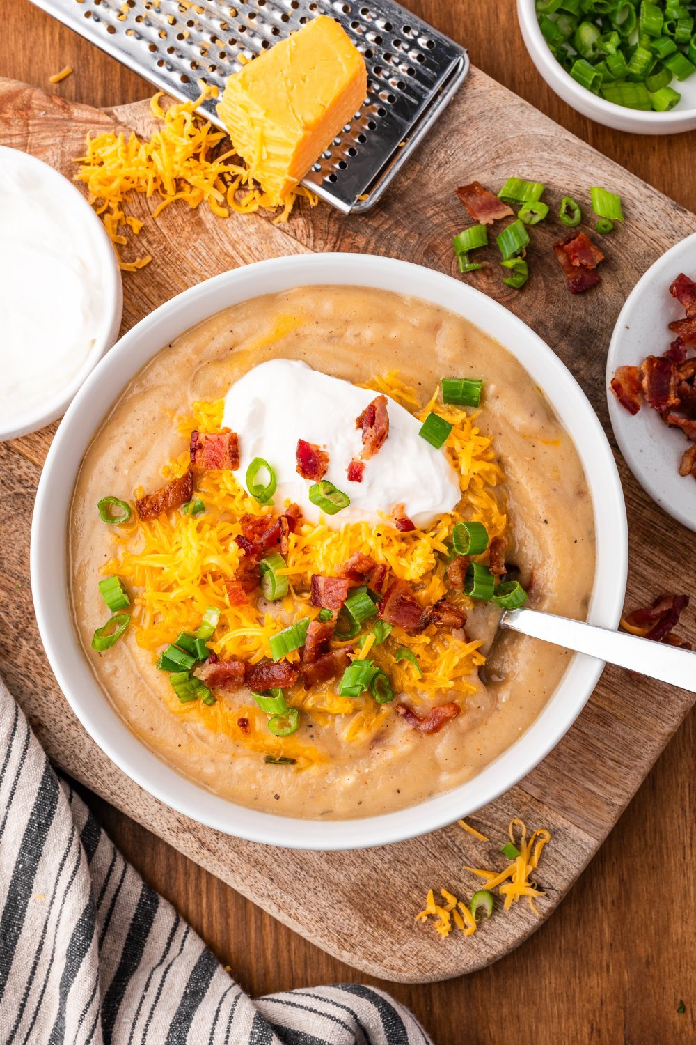 looking down at table and seeing a cutting board that is topped with a cheese grater with a block of cheese on top. A white bowl full of Crockpot Loaded Baked Potato Soup with toppings and a spoon to side. And crumbles of cheese around the bowl 
