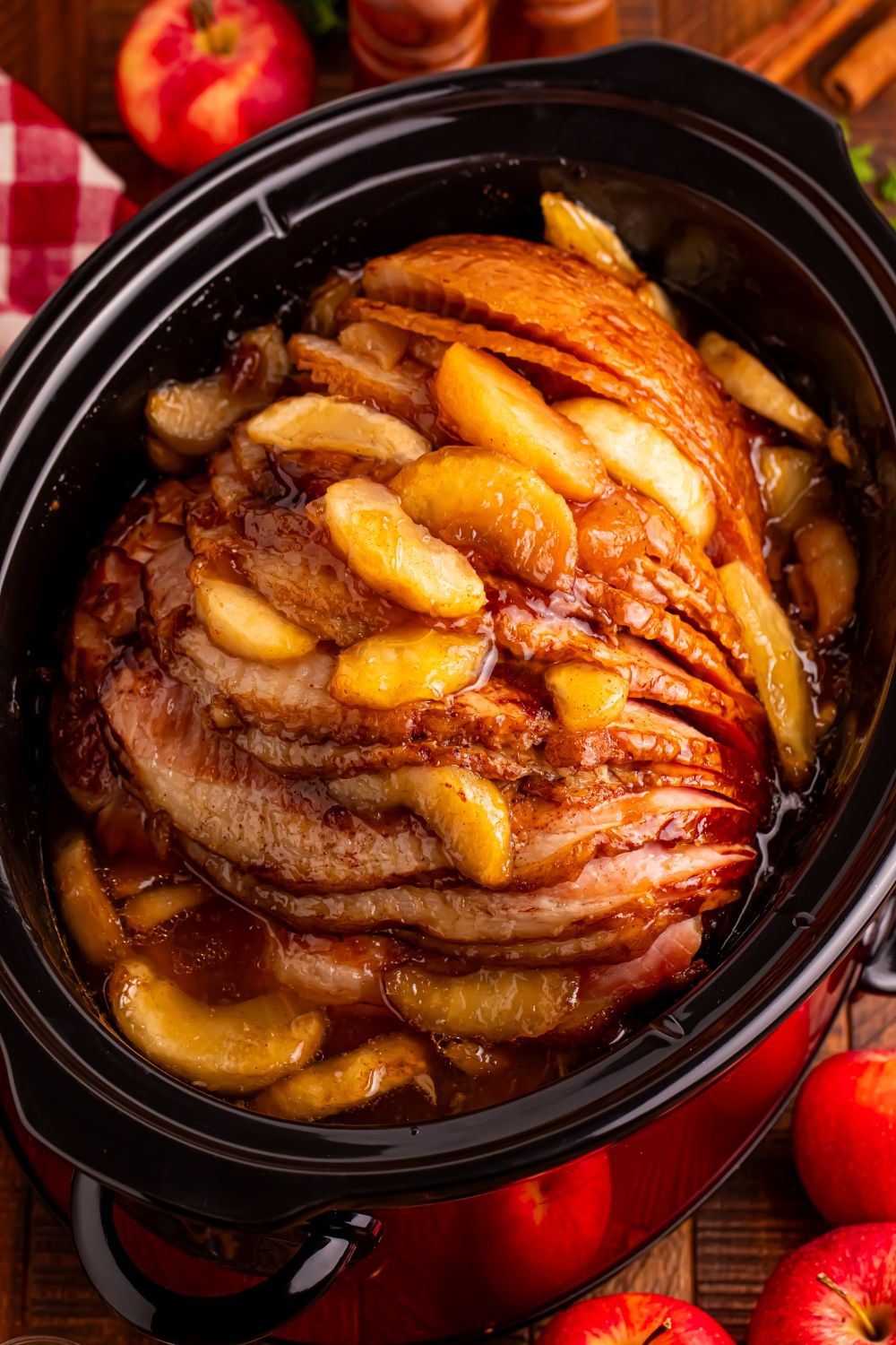 looking down at wooden table with a black slow cooker with apple glazed spiral ham. Fresh apples around table 