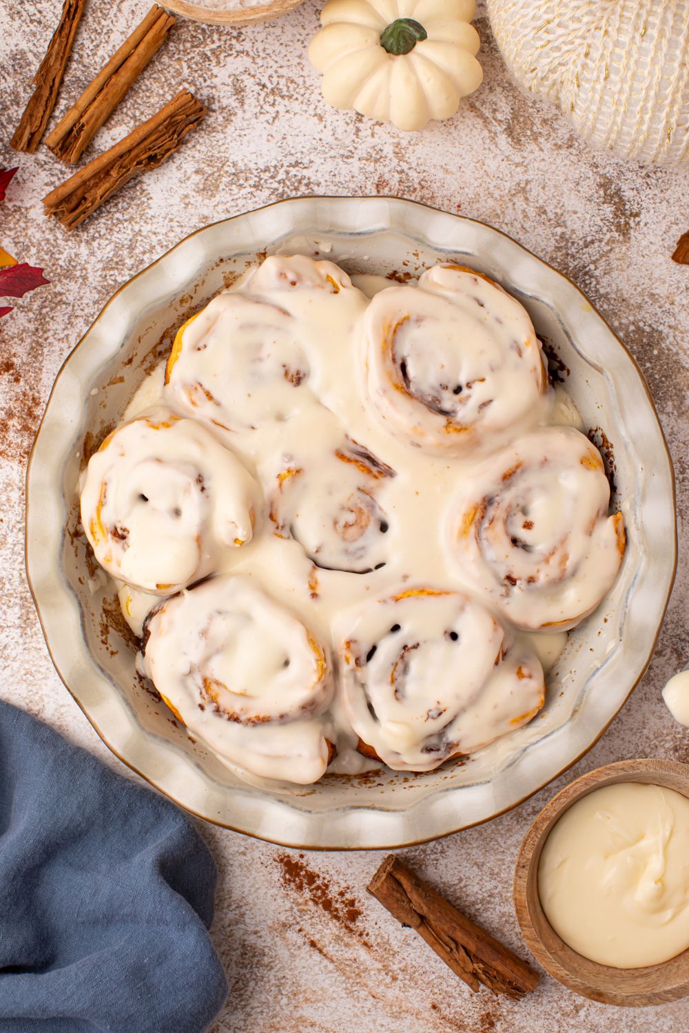 looking down to a marbled counter with cinnamon sticks, pumpkins and more around a pie plate full of pumpkin cinnamon rolls that are glazed 