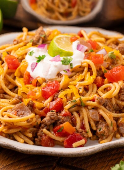 white plate on wooden table that is piled full of Cheesy Taco Spaghetti with sour cream and lime