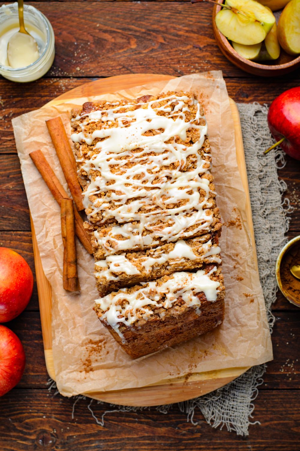 looking down on wooden table that has brown parchment paper in a rectangle with Apple Cinnamon Loaf Cake sliced and placed on it and apples placed around the table and cinnamon sticks 