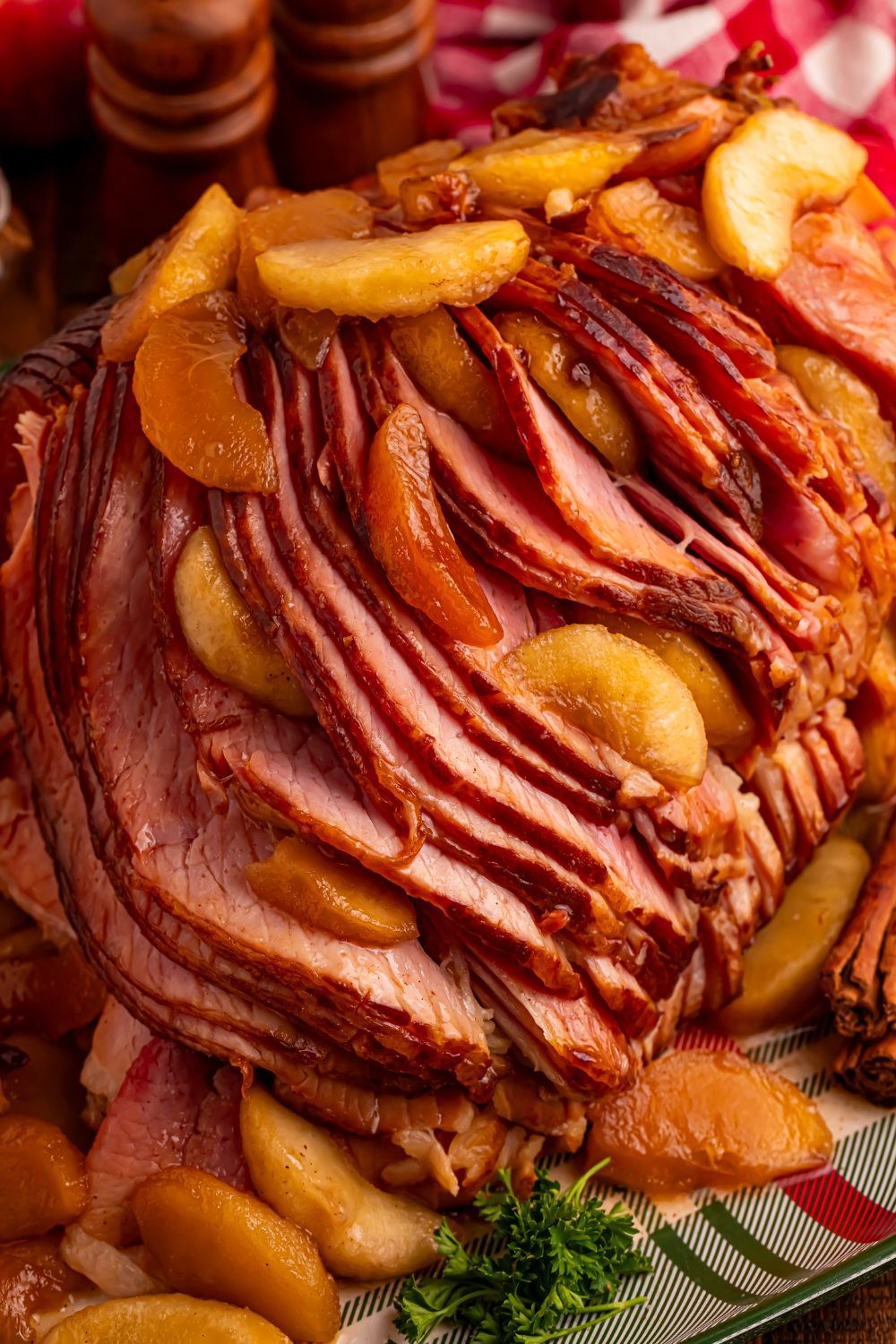 view of a large sliced spiral ham on platter with some apples on top and around with a red checkered towel behind it 