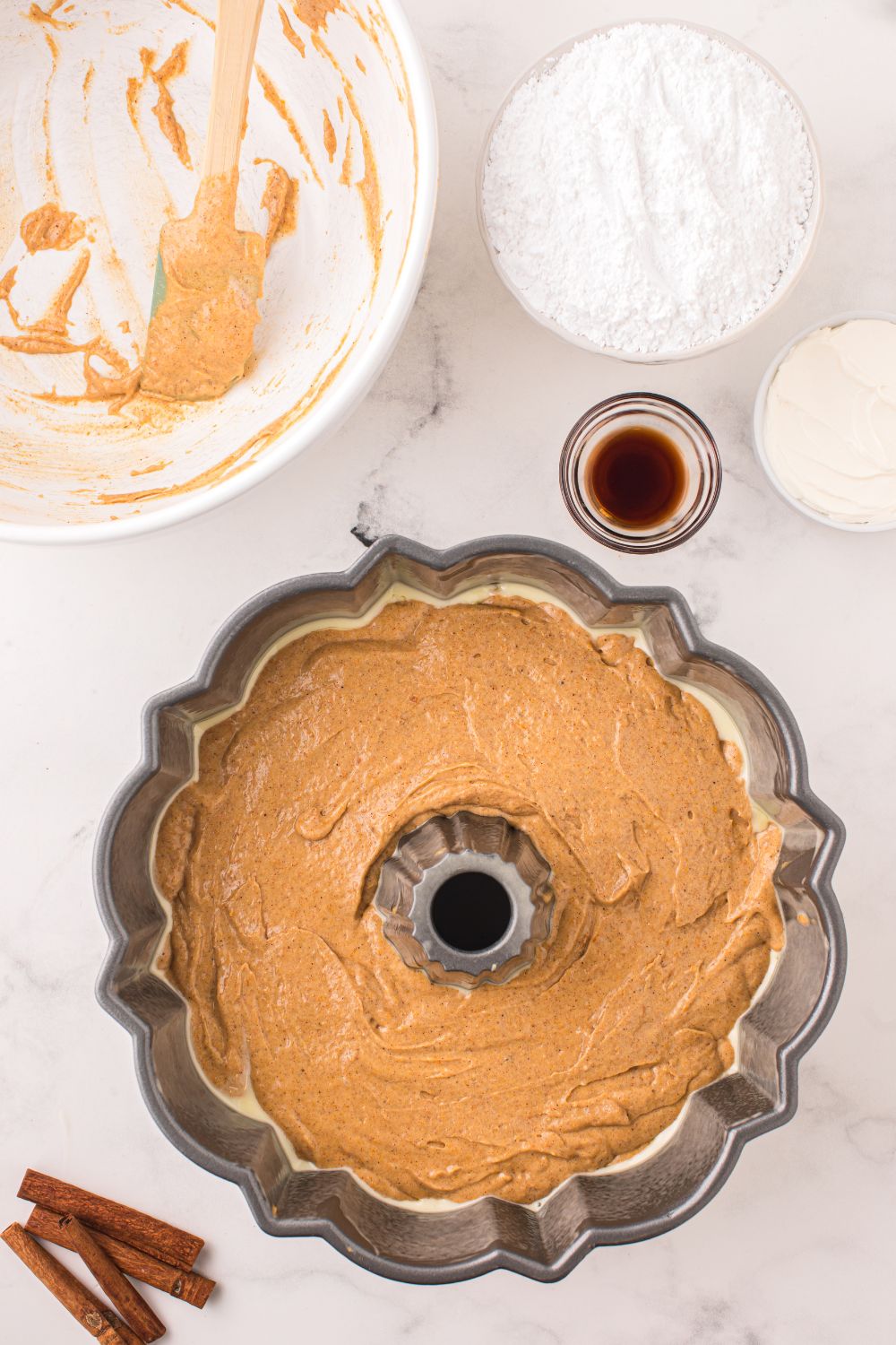 Pumpkin Bundt Cake patter in a bundt pan, with bowls around it full of ingredients for frosting 