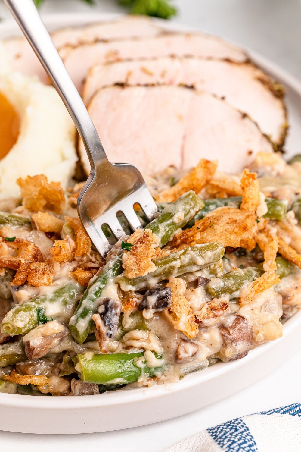 forking poking into plate full of Green Bean Casserole, with sliced turkey breast behind it and a bit of mashed potatoes and gravy