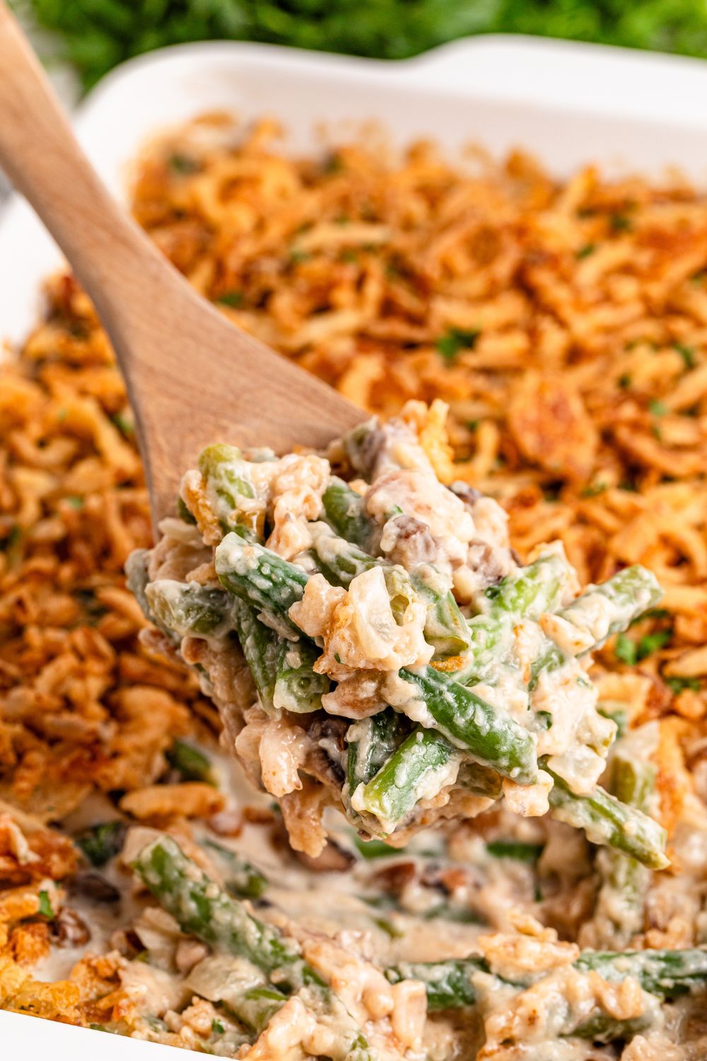 white rectangle pan on table full of Green Bean Casserole with a wooden spoon scooping some out of pan 