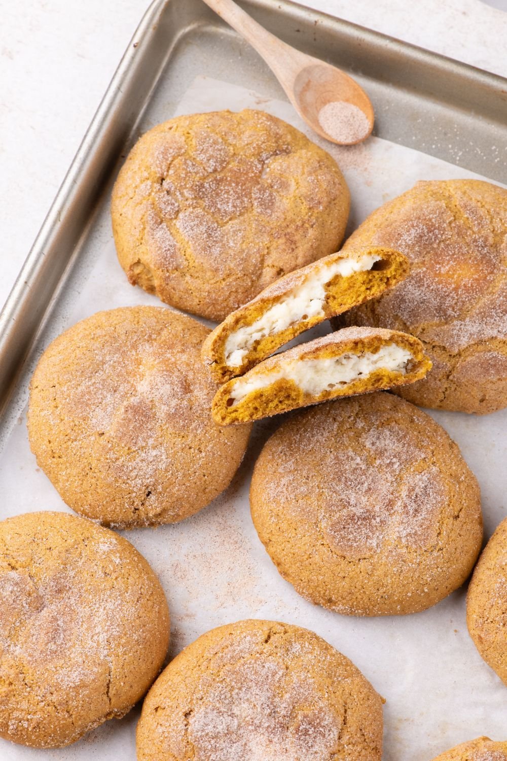 sheet pan on white table with pumpkin cookies on sheet with one cookie broken in half to see the cheesecake filling 