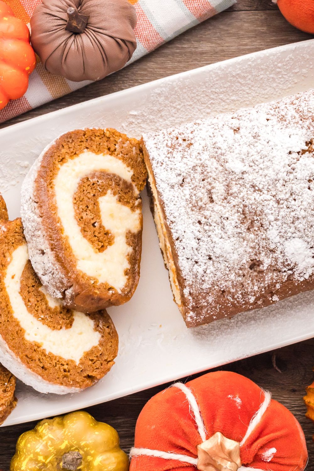 Pumpkin Cake Roll on a long white platter, with part of it whole and the other part sliced. On the wooden table around the platter are mini pumpkins for a rustic fall display 