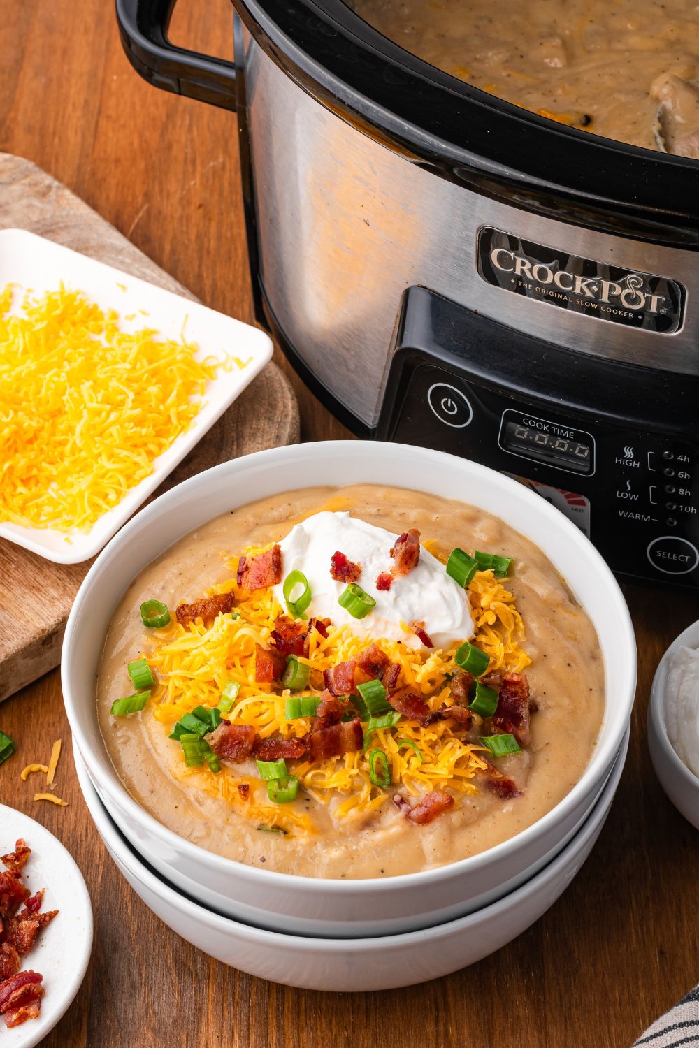 Crockpot in the background of picture. A wooden table that has two white bowls stacked and the top bowl is full of potato soup topped with shredded cheese, bacon, onions and a dollop of sour cream