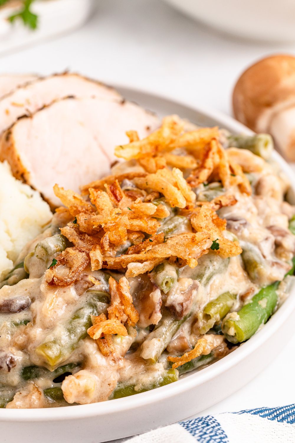 Green Bean Casserole on a round plate with some sliced turkey behind it on table and a striped towel in front of plate 