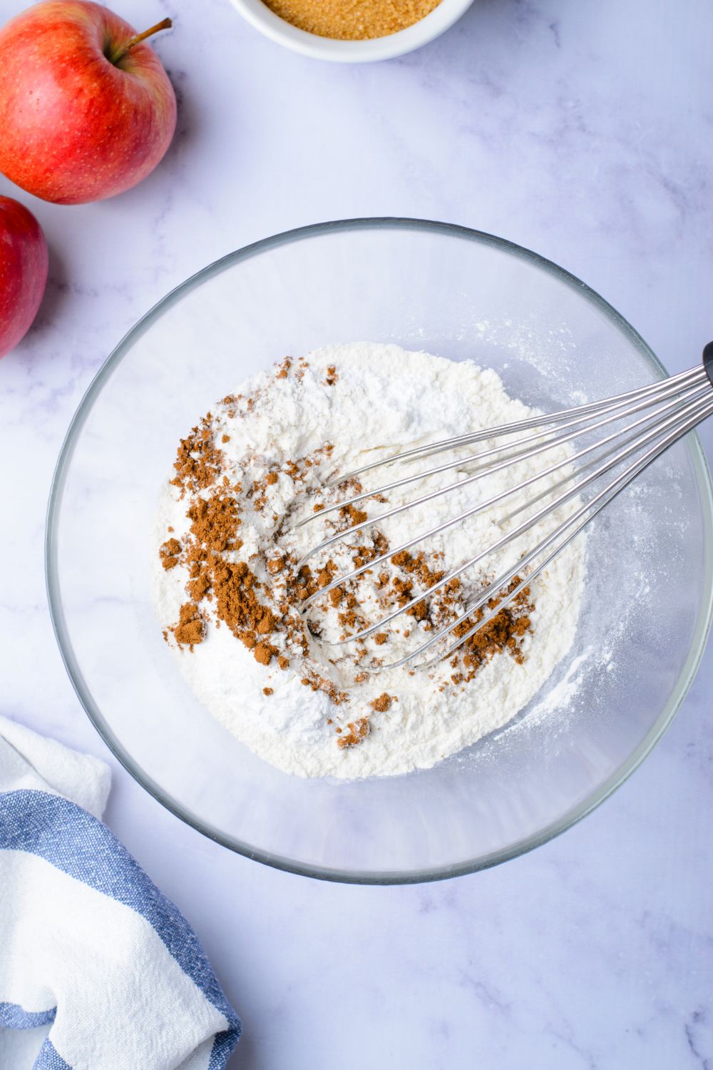flour and cinnamon mixture in a glass bowl on marble counter with two apples set to the side on table