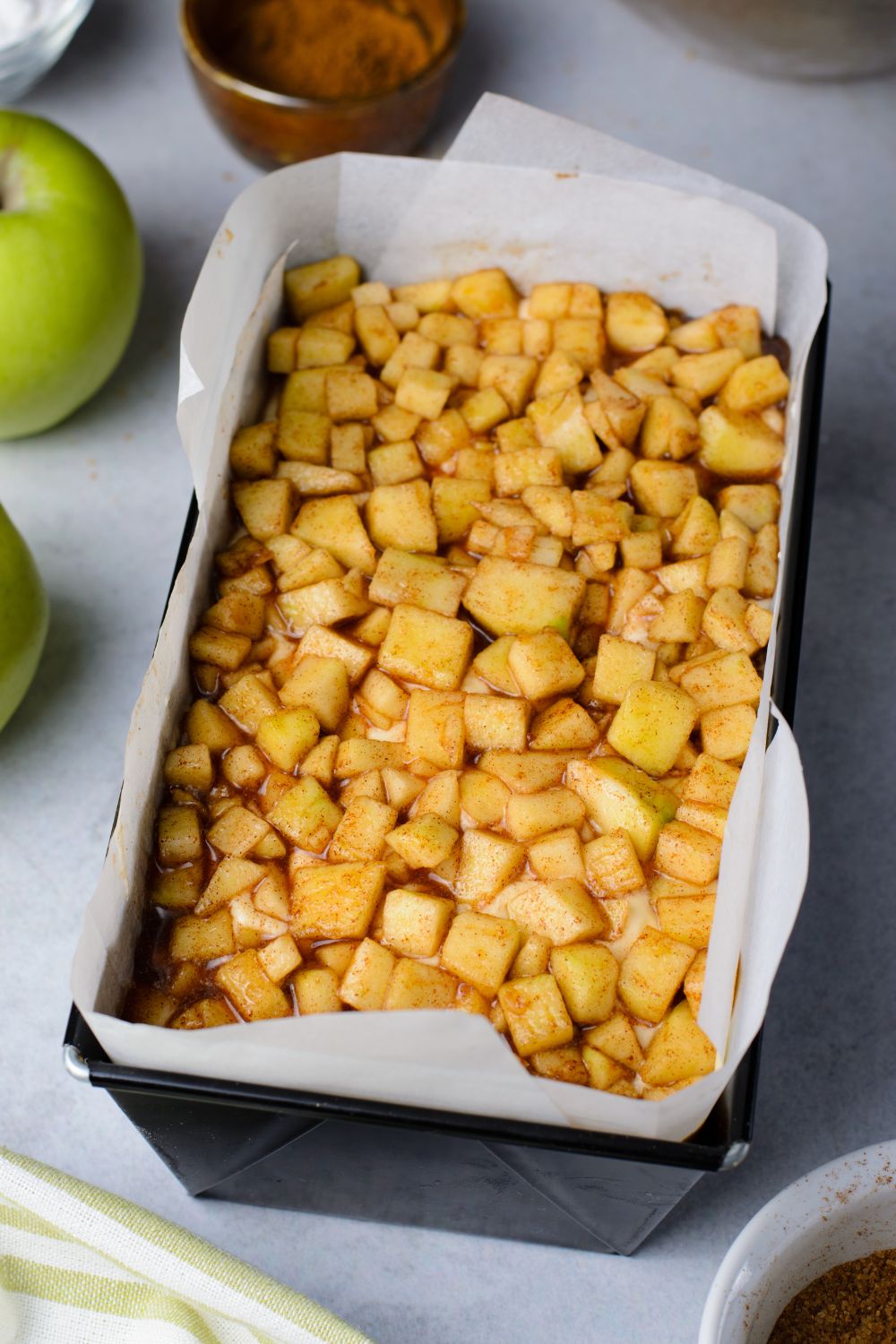 diced apples in a loaf pan for making a fritter bread with apples around the pan on table and a cinnamon mixture in a bowl