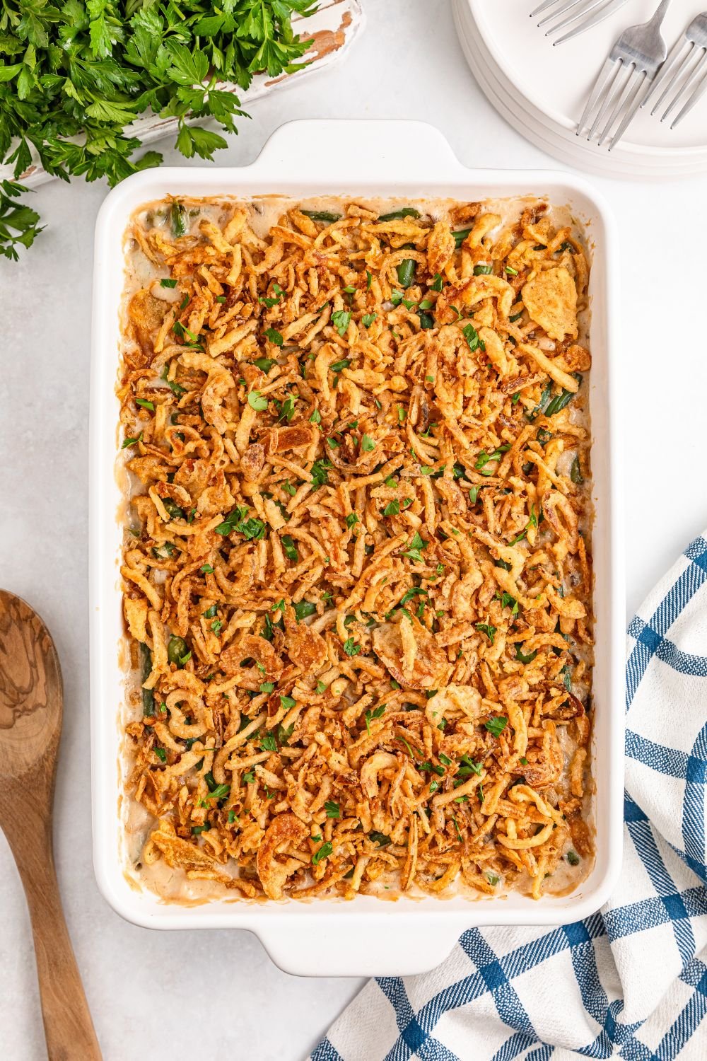 looking down on table to see a casserole dish with golden brown Green Bean Casserole and a wooden spoon to side, table napkin and fresh parsley 
