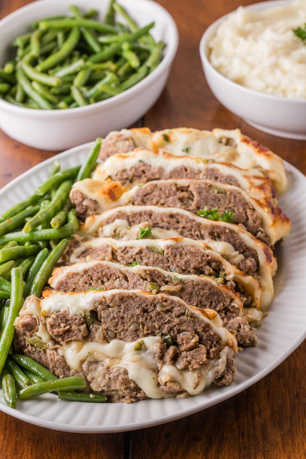 wooden table with a bowl of green beans, bowl of mashed potatoes, and a platter with sliced Stuffed Philly Cheesesteak Meatloaf on platter with green beans on side