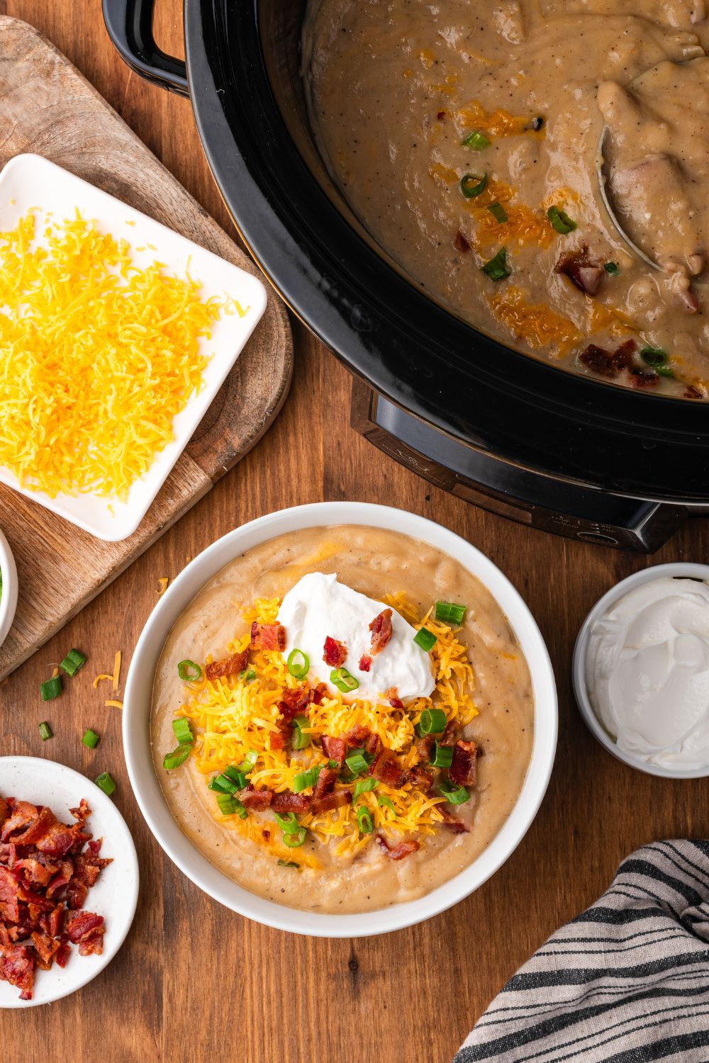 Crockpot Loaded Baked Potato Soup in a black crockpot sitting on table. Then a white bowl full of soup and toppings, a small shredded cheese, and a small bowl of bacon crumbles