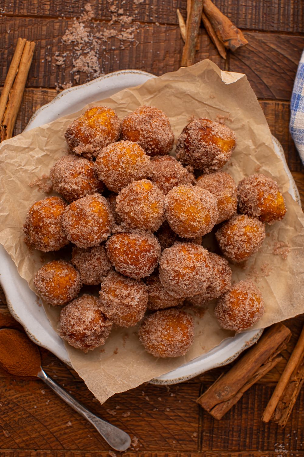 Pumpkin Pie Donut Holes on a parchment lined white platter on wooden table. There are cinnamon sticks scattered around the table 