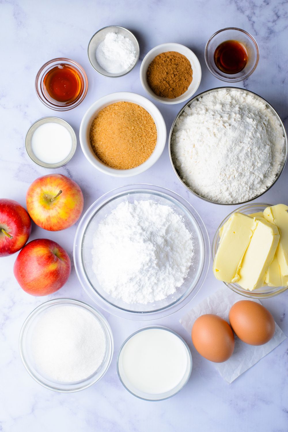 all the ingredients for Apple Cinnamon Loaf Cake in single serve small glass bowls on marble counter and whole apples and eggs included