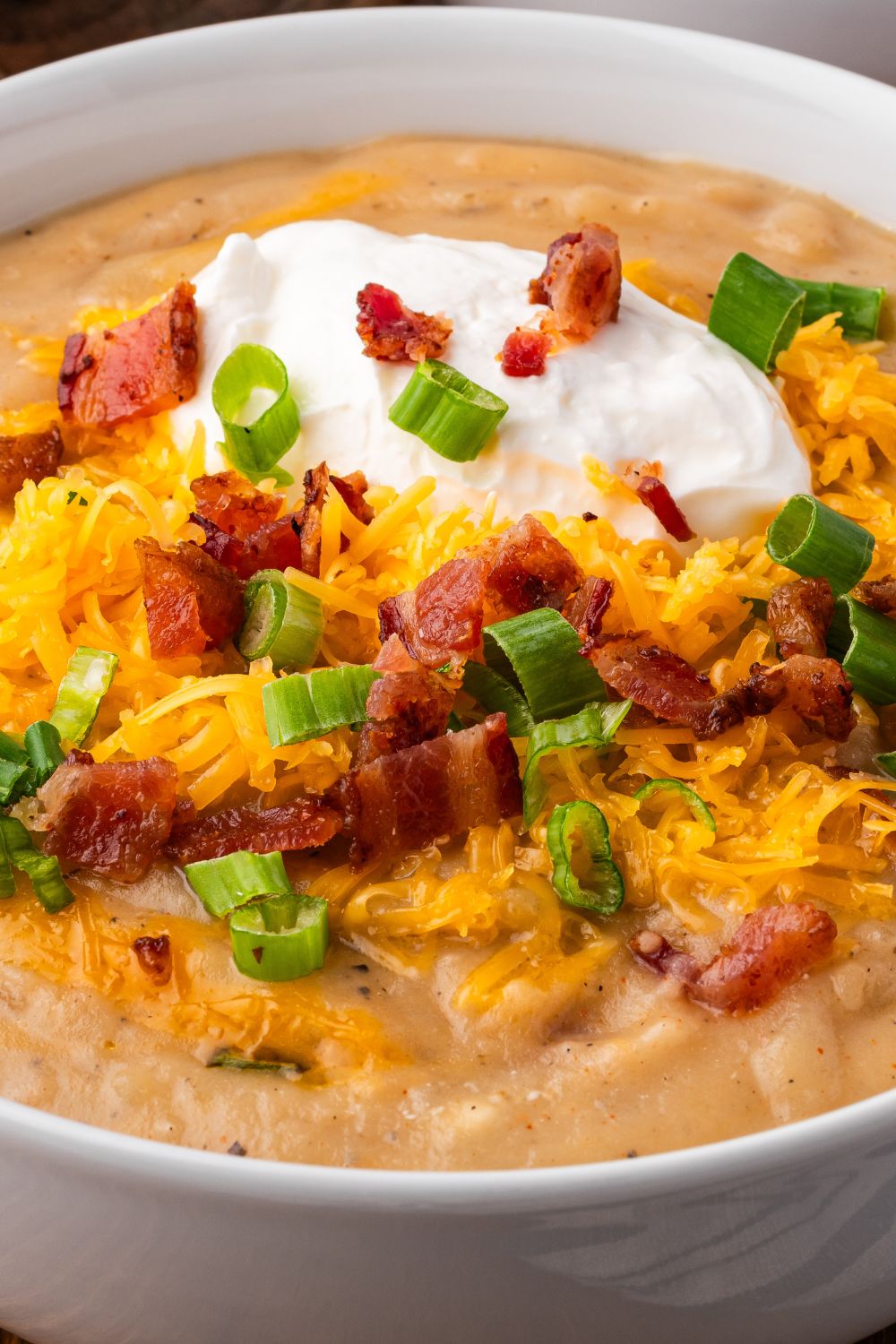 close up of Crockpot Loaded Baked Potato Soup in a white bowl on table with shredded cheese, bacon, and green onions and a dollop of sour cream for garnish 