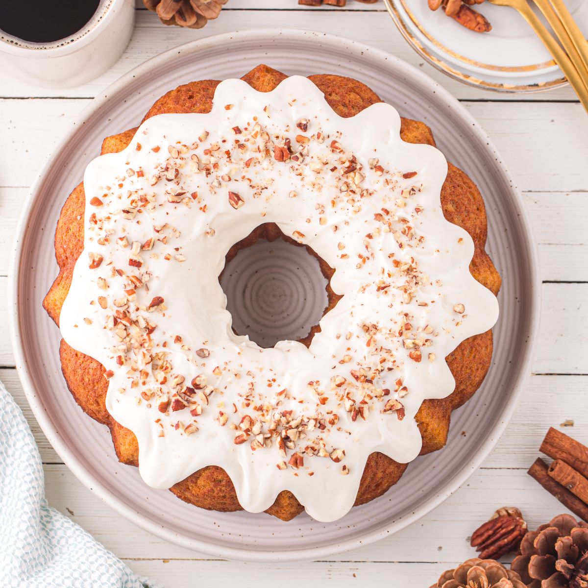 Pumpkin Bundt Cake with Cream Cheese Frosting sitting on a platter on a wooden table with cinnamon sticks and pine cone around it on table 