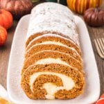Pumpkin Roll on a long platter sitting on wooden table with pumpkins around to set a fall theme. Part of the pumpkin roll is sliced and the other part is not sliced and dusted with powdered sugar