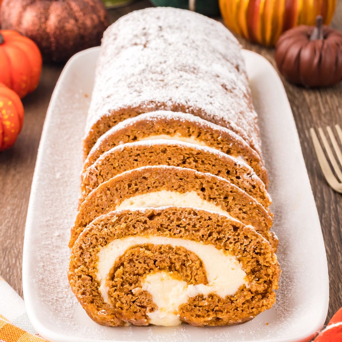 Pumpkin Roll on a long platter sitting on wooden table with pumpkins around to set a fall theme. Part of the pumpkin roll is sliced and the other part is not sliced and dusted with powdered sugar 