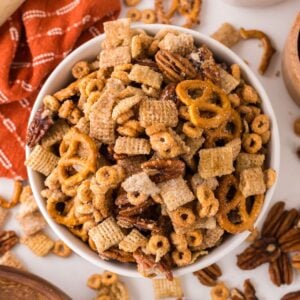 Pumpkin Spice Chex Mix in a white bowl on table with more mix scattered on table with a towel to the side of the bowl