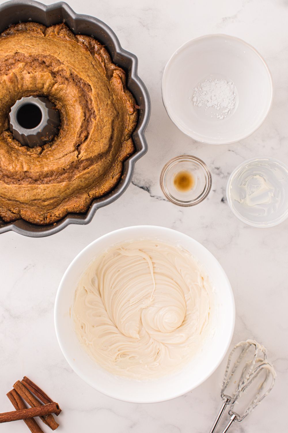 baked pumpkin bundt cake in pan with all the ingredients for cream cheese frosting in bowls around table 