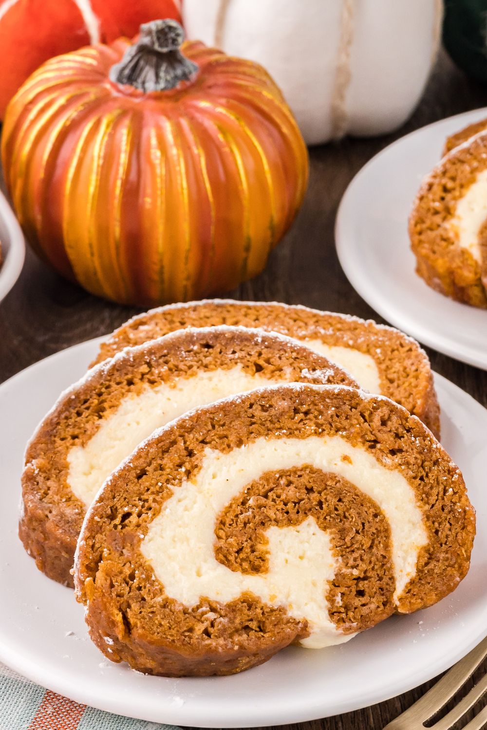 Pumpkin Cake Roll sliced on white plates placed on table with a few faux pumpkins around the plates to give a fall theme to table 