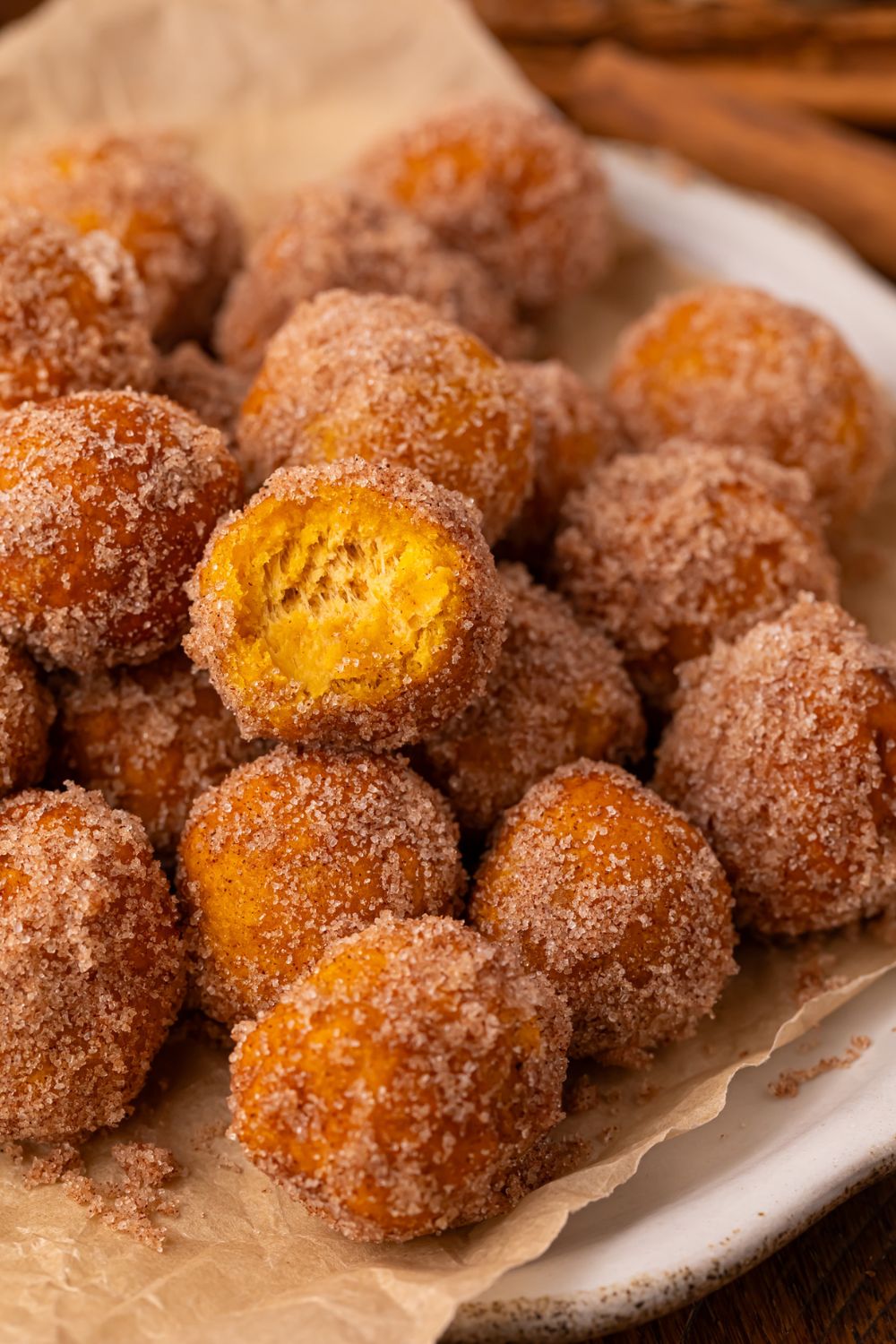 Pumpkin Pie Donut Holes scattered on a parchment lined white platter on a wooden table. The top donut hole has a bite taken out of it. 