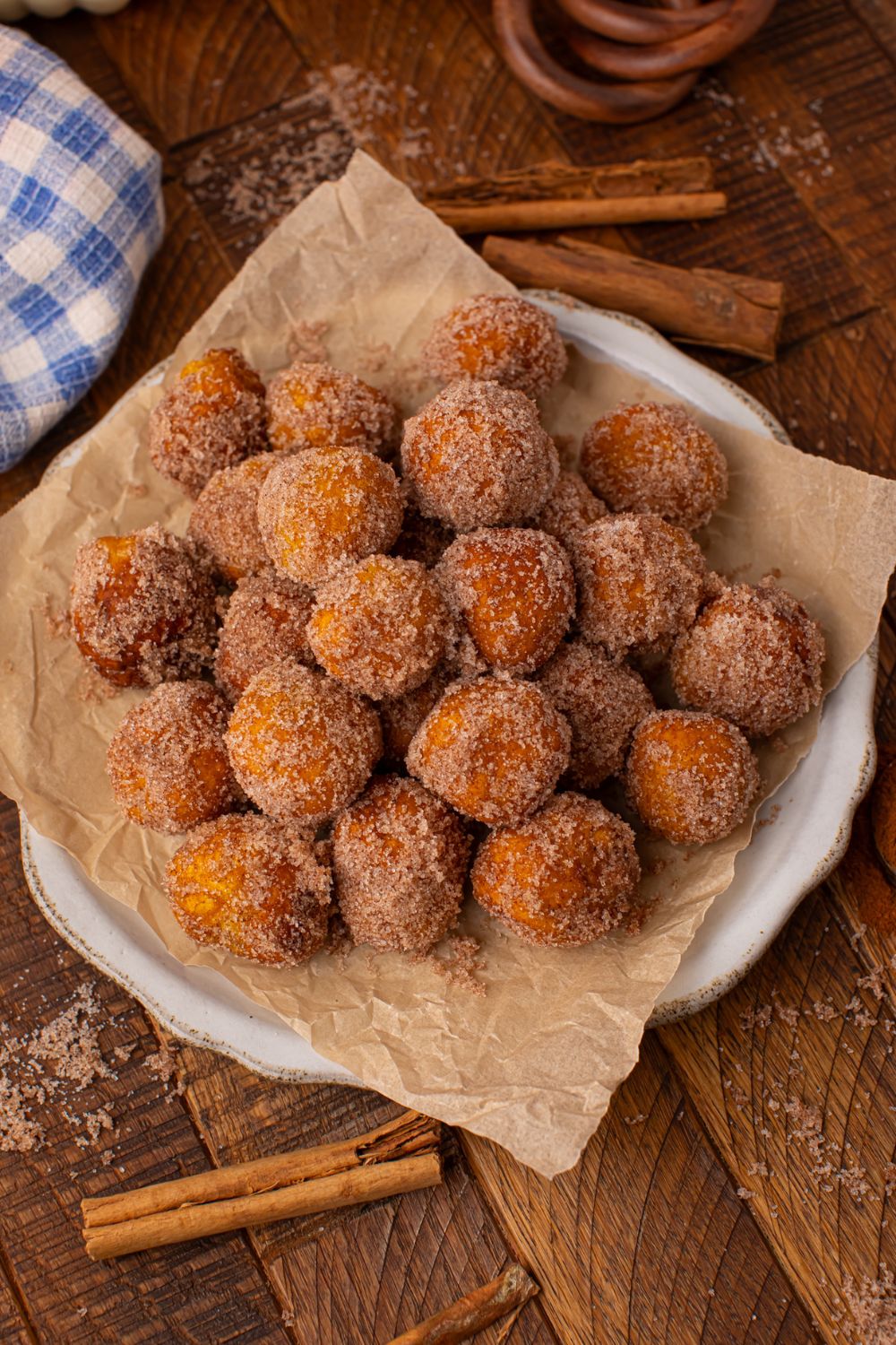 wooden table with a white platter full of Pumpkin Pie Donut Holes. Cinnamon sticks and a blue checkered towel to side of bowl