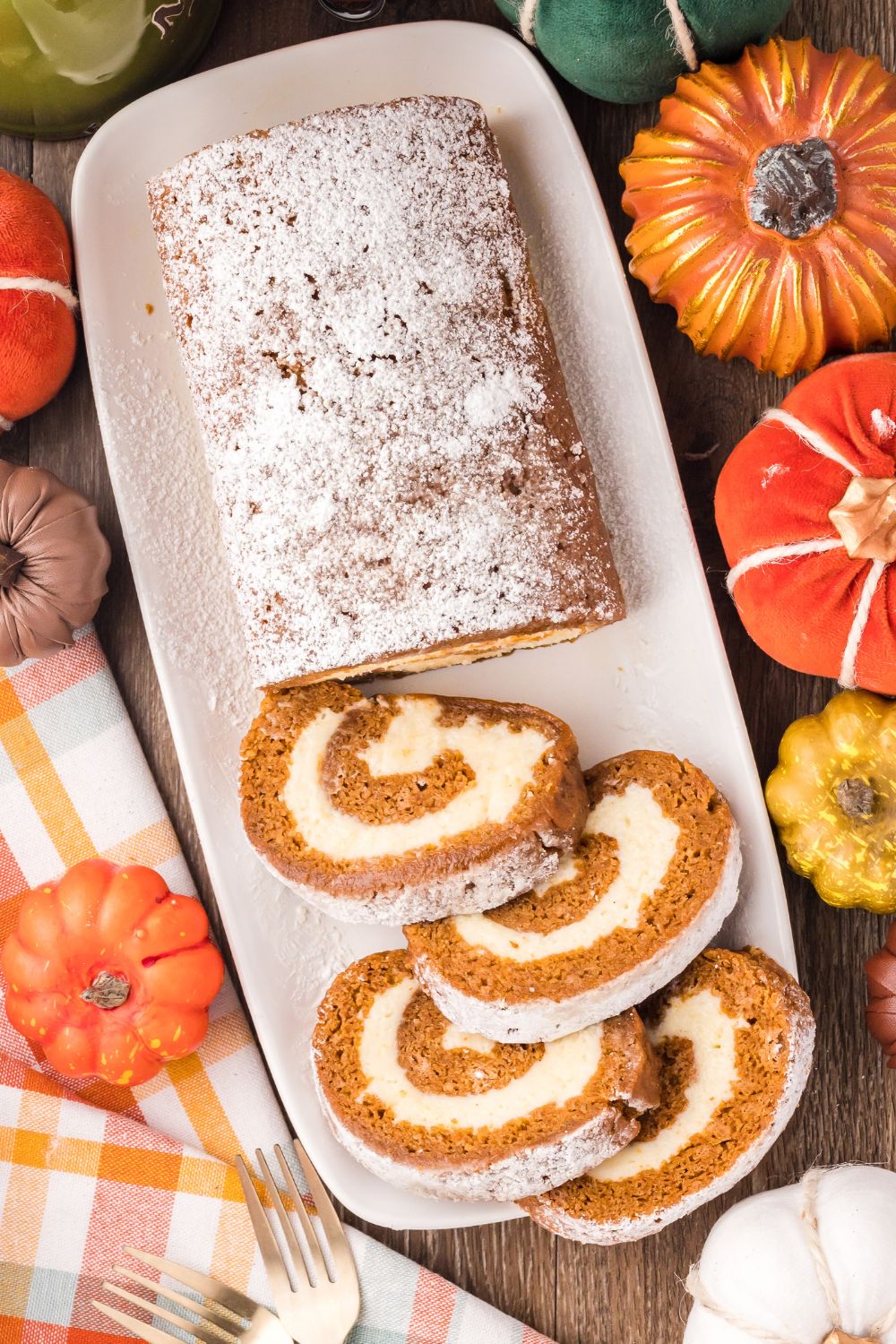 Pumpkin Cake Roll on a long white platter, with half of the pumpkin roll sliced. Faux pumpkins around the table with a white and brown striped towel