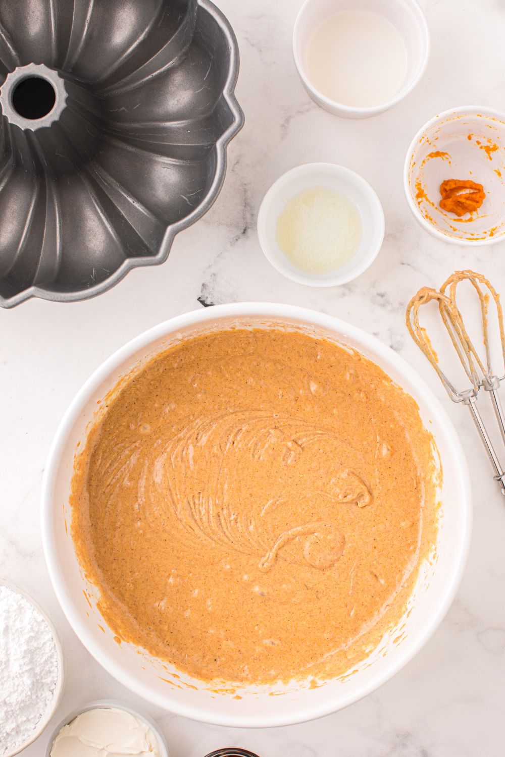 white bowl on table full of cake batter and a whisk to side and bundt pan