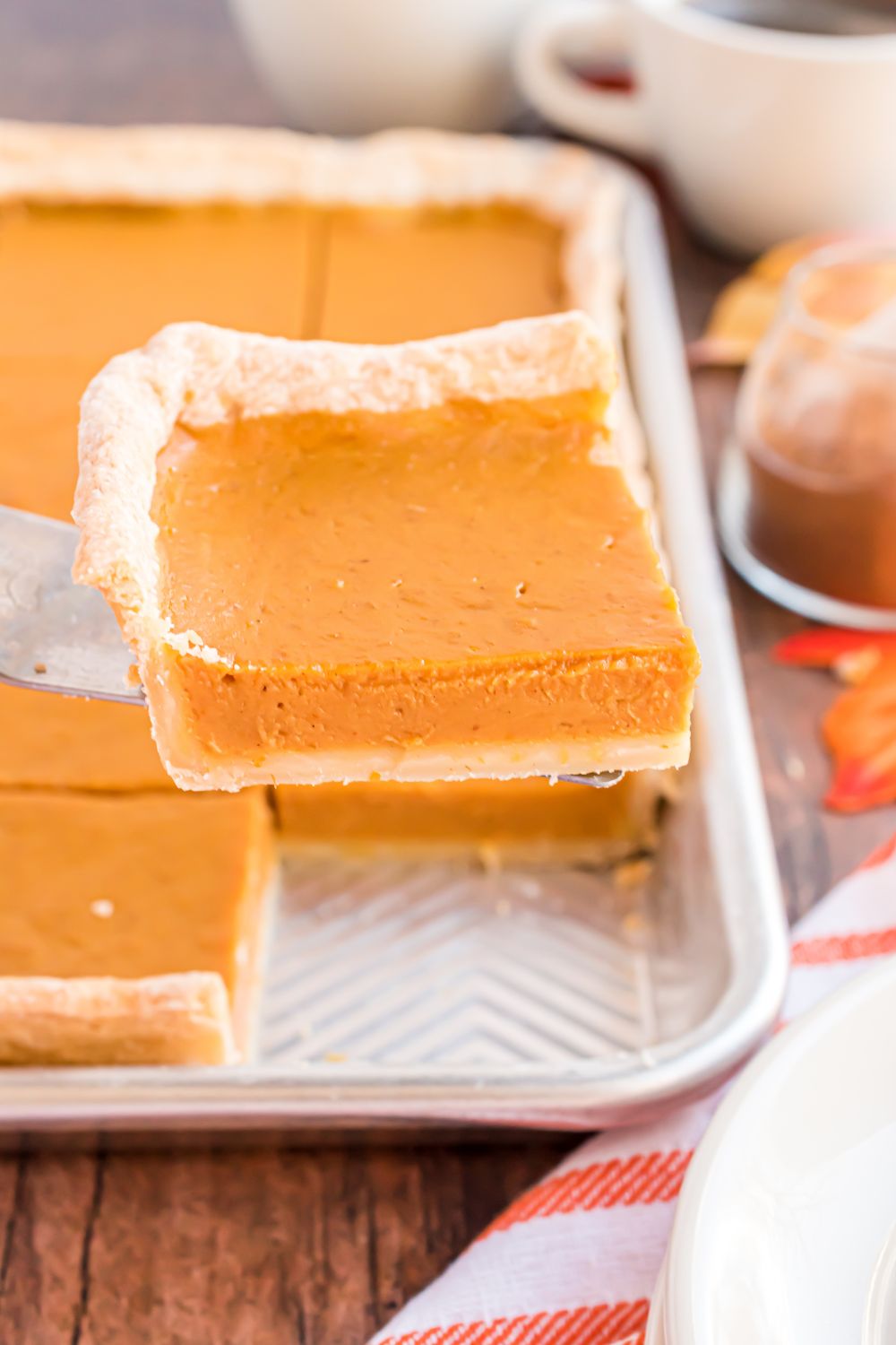 Sheet Pan Pumpkin Pie in sheet pan on wooden table decorated for fall. Once slice of the pie beign lifted out of the tray to show the thickness 